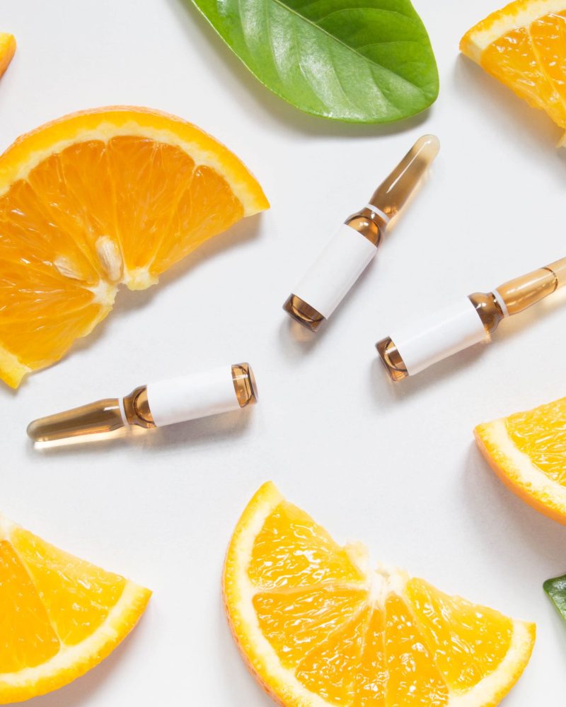 Vitamin C brown ampule for injection with fresh juicy orange fruit slices on white table. High dose vitamin c synthetic for white skin. Beauty product branding mock-up. Healthy lifestyle. Top view.